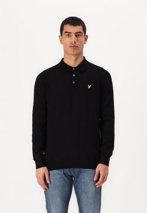Schwarzes Langarm-Polohemd aus strukturiertem Stoff mit Knopfleiste und einem gelben Adler-Logo auf der linken Brust. Getragen mit blauen Jeans.