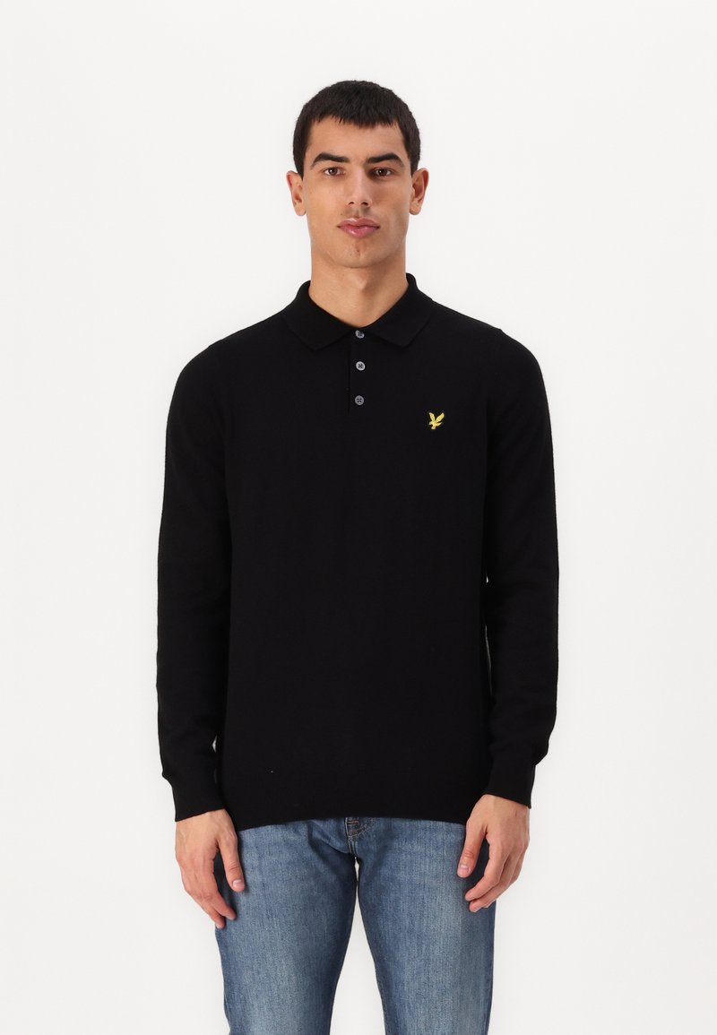 Svart långärmad polo med texturerat tyg, tre-knappsknäppning och en gul örnlogotyp på vänster bröst. Bärs med blå jeans.