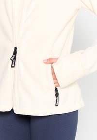 Kurtka polarowa w kolorze off-white z zamkiem z przodu, bocznymi kieszeniami i czarnymi akcentami. Charakteryzuje się gładką fakturą i dopasowanym krojem.
