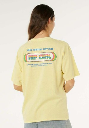 Persona con capelli lunghi castani che indossa una t-shirt giallo pallido di Rip Curl con testo colorato e design retrò sul retro, in piedi contro uno sfondo bianco.