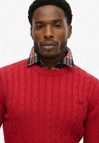 Czerwony sweter w warkoczowy splot z teksturowanej tkaniny, z koszulą w kratkę, którą widać spod spodu, podkreśloną małym logo.