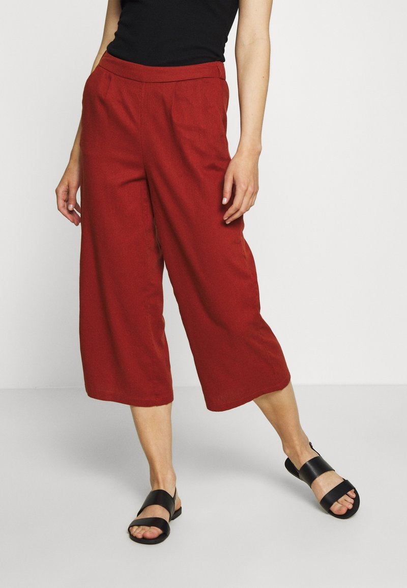 Femme portant un pantalon rouge ample et court, un haut noir sans manches et des sandales noires, debout devant un fond clair uni.