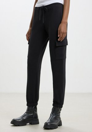 Pantaloni cargo neri con vita regolabile tramite coulisse, polsini elasticizzati e tasche laterali. Realizzati in tessuto morbido, con una superficie liscia.