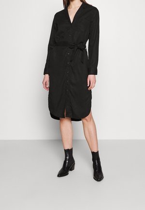 Vrouw die een zwarte, langarmige jurk met knopen draagt, met een ceintuur in de taille en zwarte enkellaarsjes, staand tegen een effen lichte achtergrond.