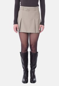 Gonna plissettata color khaki con passanti per cintura e una cintura in tessuto, indossata sopra collant neri e stivali alti neri in pelle. Texture liscia, vestibilità sartoriale.