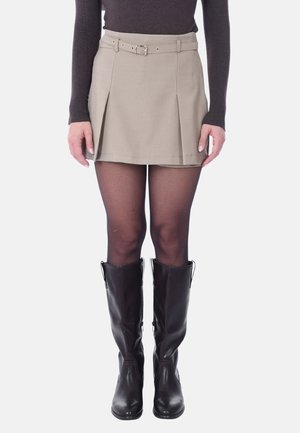 Gonna plissettata color khaki con passanti per cintura e una cintura in tessuto, indossata sopra collant neri e stivali alti neri in pelle. Texture liscia, vestibilità sartoriale.