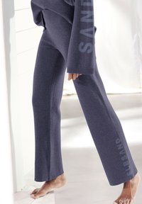 Graue, ausgestellt geschnittene Sweatpants mit einer weichen Textur. Mit einem seitlichen Druck, der in einer hellen Farbe "SANDE" entlang des Beins zeigt.