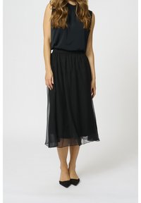 Žena v černé bezrukávové blůze, černé průsvitné midi sukni a černých špičatých balerínách, stojící před jednoduchým bílým pozadím.