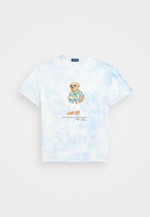 Jasnoniebieska koszulka tie-dye z nadrukiem misia w okularach przeciwsłonecznych, tropikalnej koszuli, białych spodniach i brązowych butach na środku.