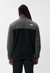 Fleecejakke i grå med en sort nederste del. Høj krave, tekstureret stof og logo på ryggen. Standard fit, lange ærmer.