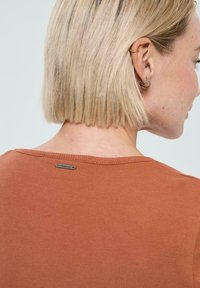 Haut côtelé de couleur rouille avec un col rond et une petite étiquette en métal à l'arrière. Texture lisse et design ajusté.