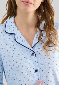 Mujer vistiendo una camiseta de pijama azul claro con un pequeño patrón floral en azul oscuro y ribete azul oscuro en el cuello y los botones.