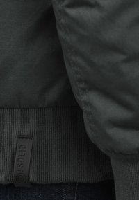 Tamnosivi kaput s kvadratnom teksturom, s rebrastim rubom i malom crnom logo oznakom. Jednostavan dizajn s mekim završetkom.