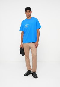 Mėlyna medvilninė marškinė su "Calvin Klein" logotipu, derinami su chaki spalvos kelnėmis ir juodais odiniais batais, laiko juodą maišą. Paprastas dizainas.