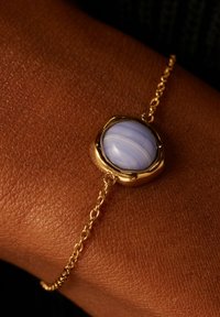 Gouden schakelarmband met een ronde lila steen, gezet in een gestructureerde zetting. De gladde afwerking van de steen contrasteert met de schakels van de armband.