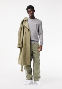 Lang olijfgroene trenchcoat, grijze trui, lichtgroene cargobroek en witte sneakers. Opmerkelijke gouden knopen op de jas.