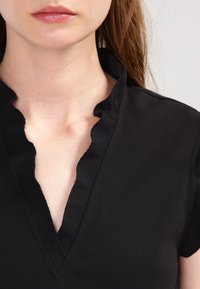 Schwarzes Oberteil mit einer gewellten Kragenlinie, kurzen Ärmeln und einem V-Ausschnitt. Glattes Material mit matter Oberfläche. Nahaufnahme des Ausschnitts.