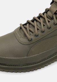 Zelené kožené tenisky s texturovaným povrchem, vybavené kulatými kovovými očky, silnými tkaničkami a drsnou gumovou podrážkou se zoubkovanými okraji.