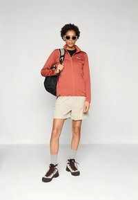 Raudona lengvai užsegama striukė iš lengvos medžiagos, derinama su smėlio spalvos trumpais šortais. Juodas kuprinė, pilkos kojinės ir rudi/juodi žygiavimo batai užbaigia įvaizdį.