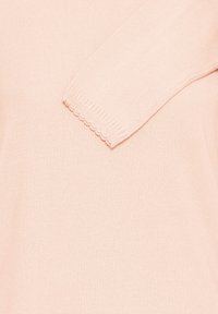 Pull rose clair en tricot présentant une texture côtelée et des bords festonnés sur les manches, offrant une finition douce et lisse.