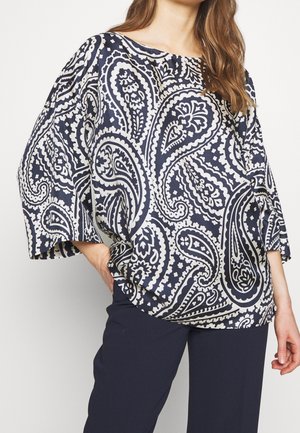 Femme portant un chemisier ample à motif cachemire bleu marine et blanc avec des manches larges et un pantalon bleu marine, debout avec une main sur la hanche.