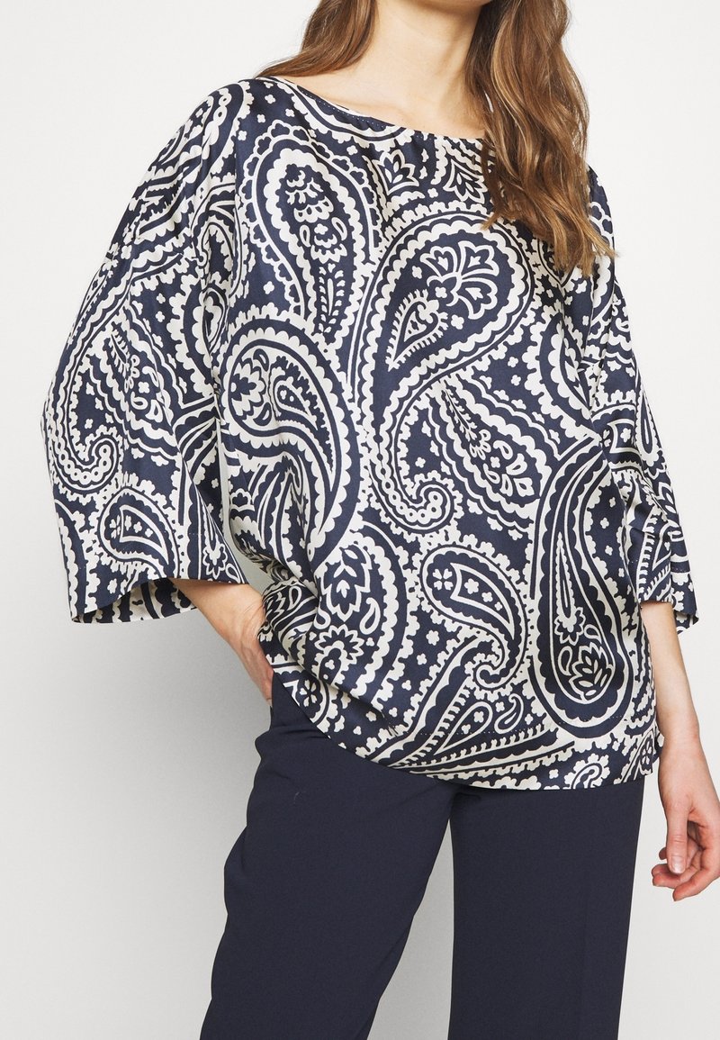 Femme portant un chemisier ample à motif cachemire bleu marine et blanc avec des manches larges et un pantalon bleu marine, debout avec une main sur la hanche.