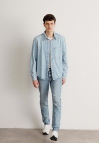 Camisa de mezclilla azul clara con un bolsillo, combinada con unos pantalones vaqueros azul claros a juego. Zapatillas blancas informales. Fondo liso. Modelo de pie.