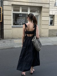 Melna, slāņaina maxi kleita ar plecu lentēm, atvērta mugura un svītraina rokassoma. Fons - pilsētas iela.