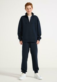 Granatowy polarowy sweter z zamkiem przy kołnierzu, w zestawie z dopasowanymi spodniami dresowymi. Materiał jest miękki, z ściągaczami przy mankietach i w pasie. Białe sneakersy.
