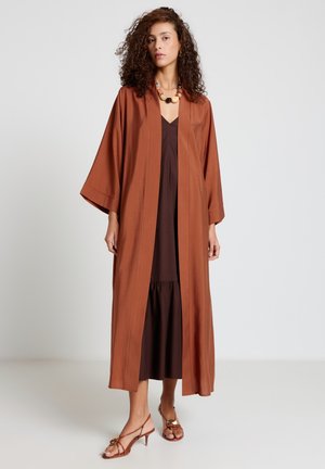 Woman with curly hair wearing a long rust-brown open coat over a dark brown dress and brown heeled sandals, standing against a plain background.