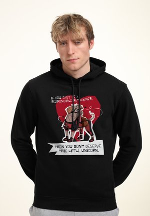 Jonge man met een zwarte hoodie met een cartoonhond in een superheldenkostuum en tekst over verantwoord huisdierenbezit.