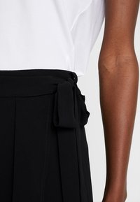 Svarta byxor i texturerat tyg med knytning i midjan. Materialet är mjukt med platta veck, och de är kombinerade med en enkel vit topp.