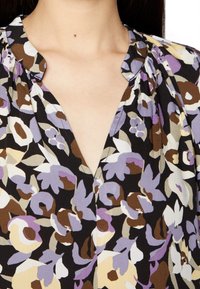 Blusa floral em preto com padrões em roxo, castanho e creme. Decote em V e ombros levemente bufantes. Tecido suave e texturizado.