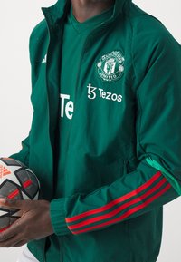 Grøn sportsjakke med røde striber langs ærmerne, Manchester United-logo og Tezos-branding. Mand holder en sort og rød mønstret fodbold.