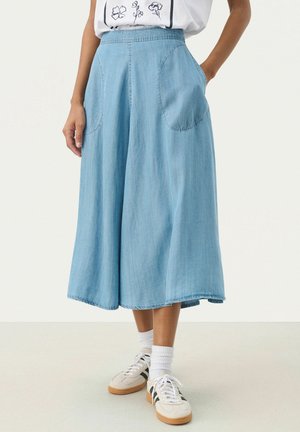 Kobieta ubrana w jasnoniebieskie, szerokie, skrócone spodnie jeansowe z przednimi kieszeniami, białe skarpetki, beżowe sneakersy z czarnymi paskami oraz białą koszulkę z nadrukiem.