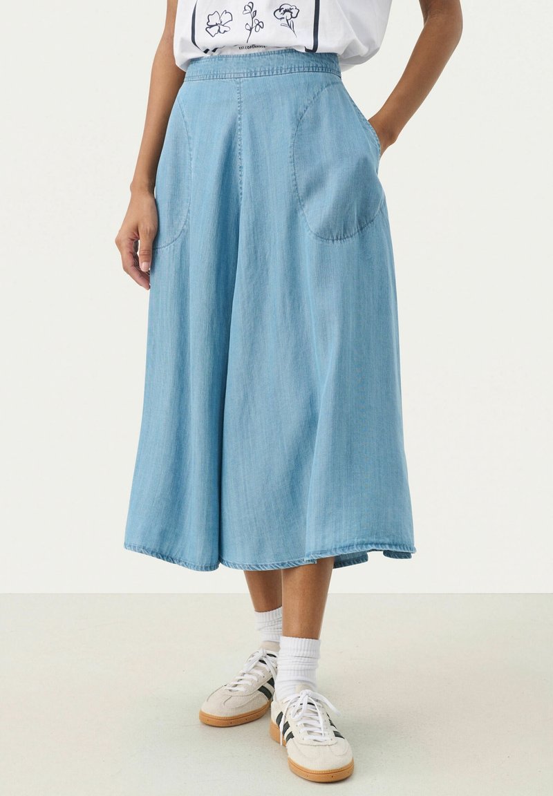 Femme portant un pantalon en jean bleu clair, coupe large et cropped, avec poches avant, chaussettes blanches, baskets beiges avec des rayures noires et un t-shirt graphique blanc.