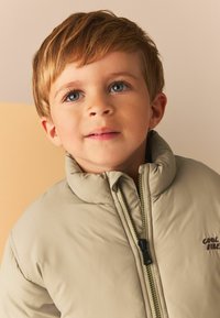 Beige pufferjacka med hög krage, dragkedjestängning och sidfickor. Har en diskret logotyp broderad på bröstet. Slät tygtextur.
