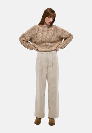 Pull en maille beige avec une coupe ample et un col montant, associé à un pantalon large beige clair avec des plis. Chaussures marron.