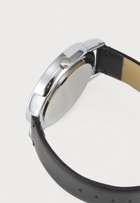 Boîtier de montre en acier inoxydable argenté de forme ronde, avec une couronne et un bracelet en cuir noir. Bracelet texturé avec des trous pour ajustement.