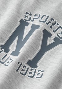 Šedá bavlnená tkanina s veľkým tmavosivým potlačou "NY" obkresleným bielou farbou, s nápisom "SPORTS" nad ním a "CLUB 1986" pod ním.