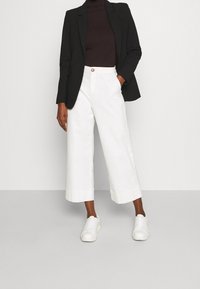 Witte, cropped broek van katoen, gecombineerd met een zwarte blazer en een donkere top. De outfit heeft eenvoudige lijnen en casual witte sneakers.