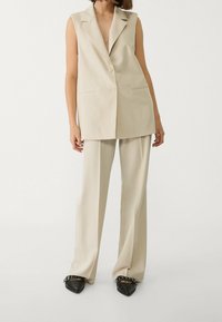 Beżowa kamizelka bez rękawów z klapami z wycięciami i dwoma guzikami, zestawiona z dopasowanymi szerokimi spodniami i czarnymi butami na szpic z ozdobami.
