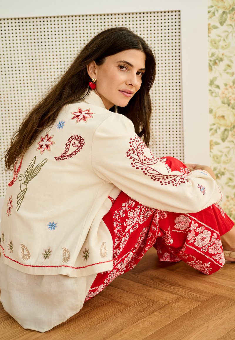 Femeie cu păr lung și închis la culoare, purtând o jachetă brodată crem și pantaloni roșii cu imprimeu floral, așezată pe podea din lemn, sprijinită de un perete decorativ.