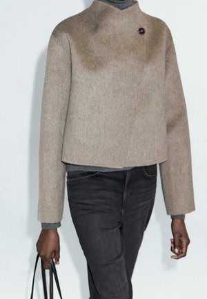 Person wearing a beige wool jacket with a single black button near the collar, dark gray jeans, and holding a black tote bag.