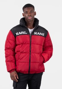 Rød puffjakke med sorte detaljer, der har høj krave, lynlås lukning og "KARL KANI" logo på brystet. Blød, quiltet tekstur.