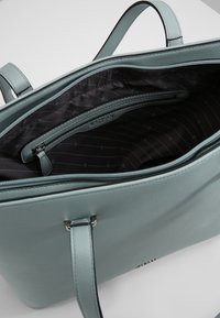 Sac fourre-tout en cuir vert d'eau clair avec deux anses. L'intérieur est doublé de tissu noir à motif rayé discret et comprend une poche zippée.