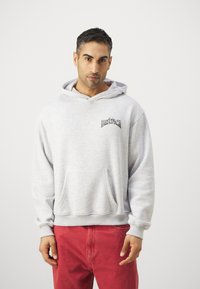 Light gray cotton hoodie with kangaroo pocket, ribbed cuffs, and hem. Features a black logo on the chest. Worn with red pants.