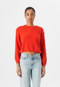Pull court rouge en tissu texturé, avec un col rond et des manches longues, associé à un jean en denim bleu clair taille haute.