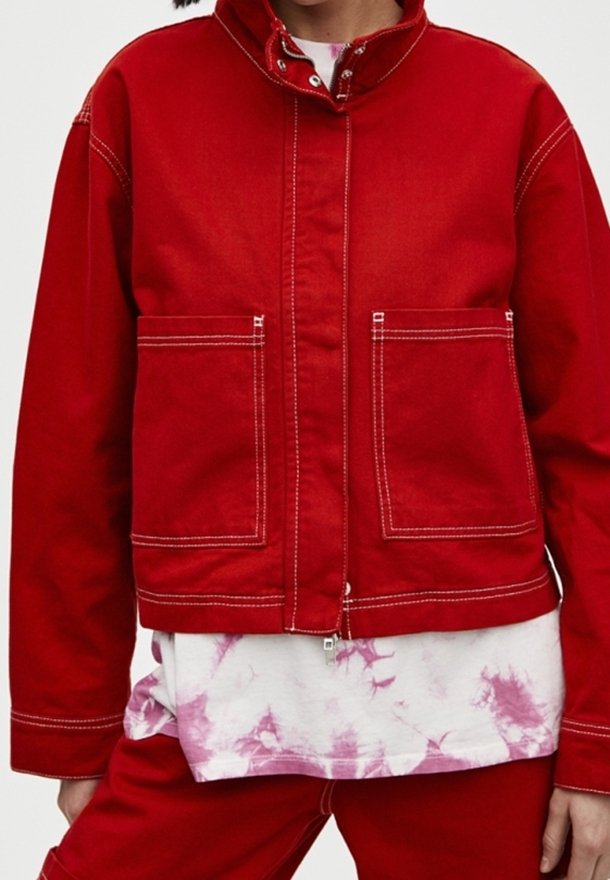 Red denim jacket with a high collar, front zip, and two large front pockets. Features white stitching and a relaxed fit. Worn over a tie-dye shirt.