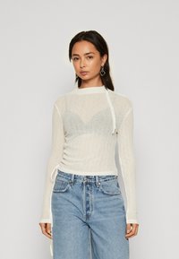 Polo alb, texturat, cu mânecă lungă, cu legături pe lateral; textură transparentă peste un sutien gri deschis. Asortat cu blugi din denim albastru cu talie înaltă.